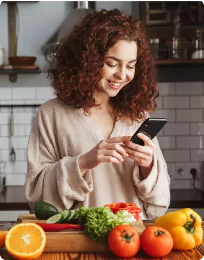 stock-photo-photo-of-joyful-caucasian-woman-holding-smartphone-while-cooking-salad-with-fresh-vegetables-in-1402613849 1.png